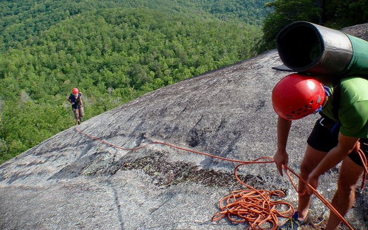 In the foreground, a person wearing safety gear is secured by ropes as they look down a rock incline at another person wearing safety gear, making their way up. On the ground below is a thick, green wooded area.